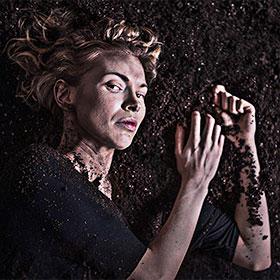 An actor lays on her side in a pile of dirt and looks at the camera. 