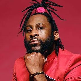 A man wearing his dreadlocks in a ponytail atop his head rests his chin on his fist and looks at the camera.