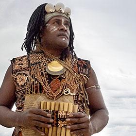 A musician wearing indigenous clothing holds a wind instrument while looking out at the ocean.