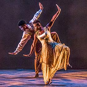 Two Black dancers wearing loose clothing pose in unison with both arms outstretched.