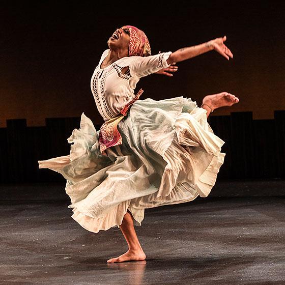 A Black woman wearing a flowing dress kicks back one leg and throws back both arms as she dances.