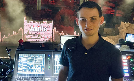 Alex Pregel stands in front of a lighting and sound control board while a production of Annie appears on stage in the background.