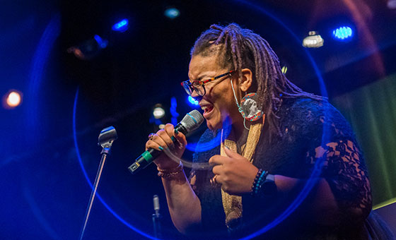 A Black woman holds the microphone to her face as she passionately sings with her eyes shut.