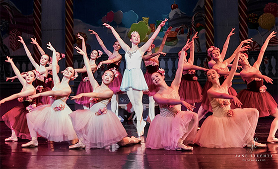 Fourteen young dancers with their arms in the air encircle a central dancer on pointe.