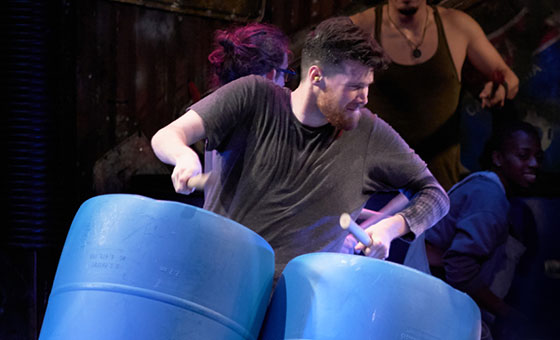 A bearded man with drum sticks pounds two large plastic barrels.