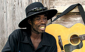 Brushy One String wears a large brimmed hat and sits in front of his guitar with one string.