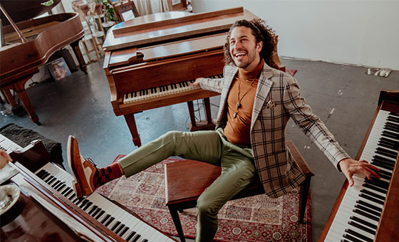 Emmett Cohen sits on a bench. His arms and legs are playing 3 pianos around him.