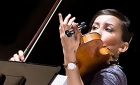 A close photo of a young Latino woman playing the violin on a dark stage.
