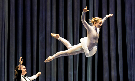 A woman in a white costume and tutu flies through the air with her arms outstretched.