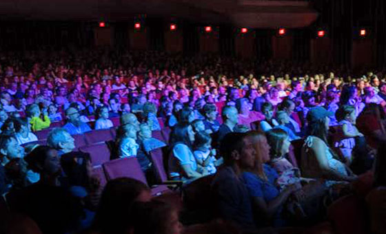 A crowd of parents with young children sit in an illuminated auditorium.