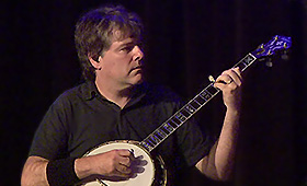 A musician watches his left hand on the fret as he plays his banjo.