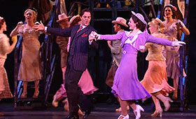 An actress depicting main character Peggy Sawyer is lead by a male character through a street full of dancing cast members.