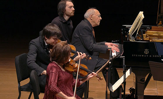 A man sits behind a pianist at his instrument while two violinists play while seated next to them.
