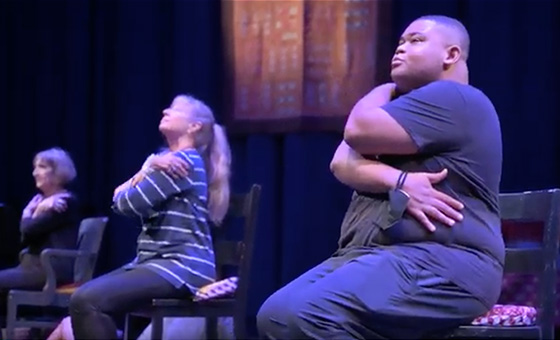 Pheolyn Allen sits in the first chair of a row on stage with his arms wrapped around his body.