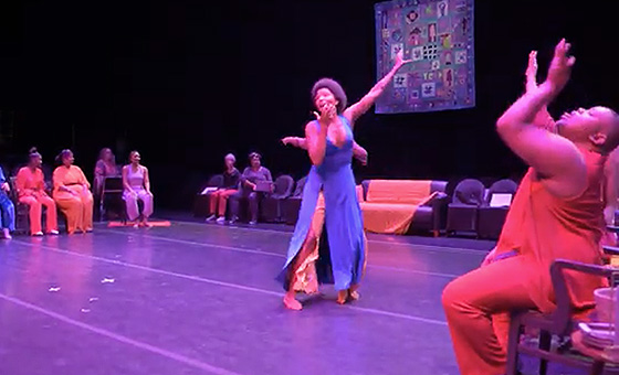 A Black dancer in free flowing clothing dances on stage. 8 other artists dance from their seats placed around the perimeter of the stage.