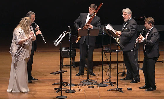 Five formally dressed musicians stand in front of music stands and play their wind instruments.