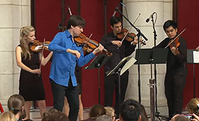 A group of 4 musicians playing stringed instruments crowd around two music stands.
