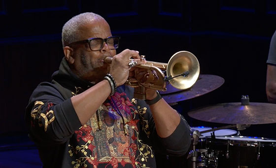 A Black man wearing eyeglasses closes his eyes as he plays a trumpet.