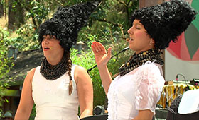 Two female members, dressed in traditional Ukrainian wedding gowns and headgear, play drums and sing into tiny microphones attached at their ears.