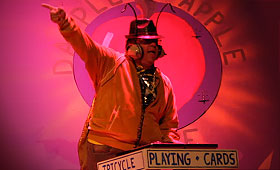 An actor portrays an insect dressed as a club DJ and points his finger upward.