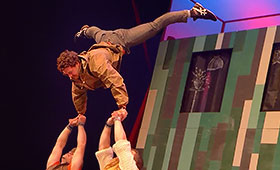 An acrobat does a handstand on the outstretched arms of two men below him.