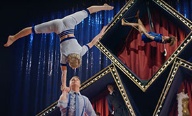 A male acrobat holds another’s hands while she straightens her body into an upside-down split position.