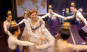 Children hold hands and move in a circle around a woman wearing a Victorian-age dress.