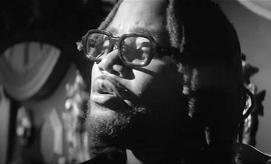 A close up photo of Leon Thomas's head as he sings in the room of an extravagant house.