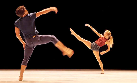 Two dancers reach their right arms and legs out in front of them in a high lunging motion.