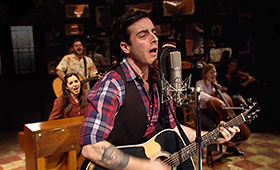 Guy, the main character in Once, sings into a microphone and plays guitar while musicians and members of the cast in the background assist in the musical performance.