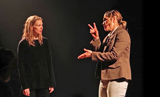 A woman stands and watches another woman making sign-language gestures with her hands.