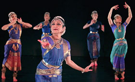 A woman reaches forward while four women dance behind her, all wearing traditional Indian clothing.