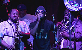 Three musicians play the saxophone, tuba, and drums behind a man singing in sun glasses on stage.