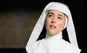 A nun wearing a habit in a dark room looks up while singing.