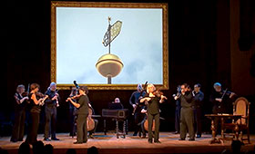 Members of Tafelmusik Baroque Orchestra stand on a stage below an oversized framed picture of a building spire while they perform on their classical instruments.