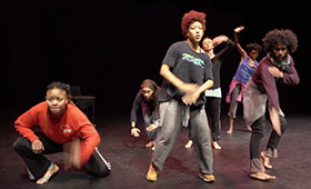 Dancers wearing baggy clothing kneel and make hand and arm gestures.