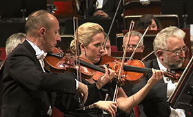 A photo highlights violinists dressed in formal attire performing with the Warsaw Philharmonic Orchestra.
