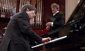 Pianist Seong-Jin Cho plays the piano while Warsaw Philharmonic Orchestra Music and Artistic Director Jacek Kaspszyk watches him from the right.