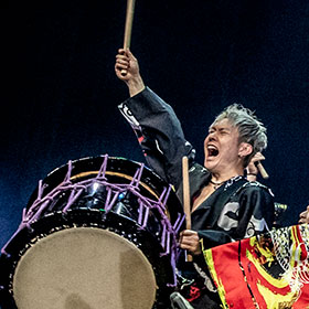 A Japanese man holding a wadaiko drum and drumsticks raises one arm and yells out.