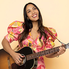 A woman with long straight hair smiles while she plays an acoustic guitar. 