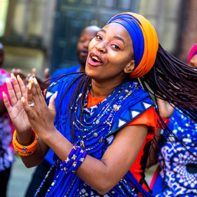 A woman wearing a headscarf and boldly patterned clothing claps her hands and sings.