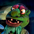 A green giant puppet with a large nose stands in front of the moon.