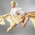 A female dancer holds on to the folds in her skirt at each hip as she kicks her right leg straight and bends her left leg up while pointing her toes.
