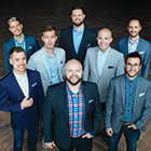 Eight male singers wearing sport jackets and button-up shirts stand in a huddle and smile at the photographer.