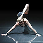 A contortionist poses with both hands on the ground in front of her, one leg outstretched behind and the other leg bent up and over to touch her head.