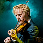 A man plays his violin while a light haze surrounds him on stage.