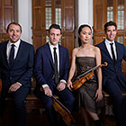 A quartet—three men wearing suits and a woman holding a violin and dressed in a simple formal dress—sit for the camera.