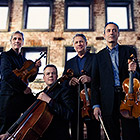 The quartet—violinists Drucker and Setzer, violist Dutton, and cellist Watkins—pose with their instruments in front of a building.