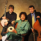 A banjo player and a double bassist stand and hold their instruments while a man sits between them and smiles.