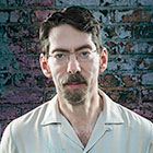 Jazz bandleader and musician Fred Hersch, standing against a distressed brick wall, looks directly at the camera in a portrait shot.